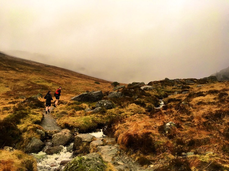 Two runners crossing a stream in sodden grass and cloud